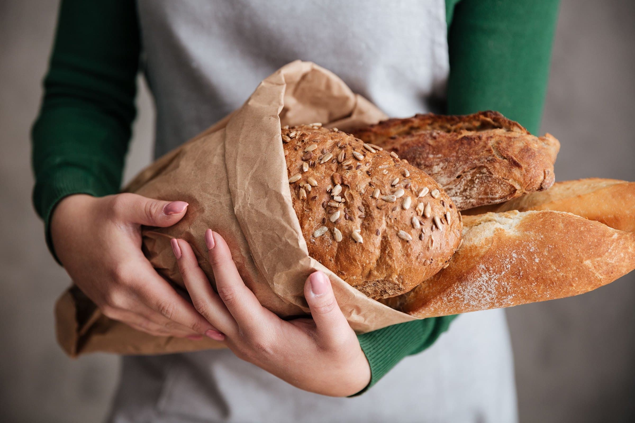 Hands in a green sweater cradling a paper bag of seeded loaves and a baguette