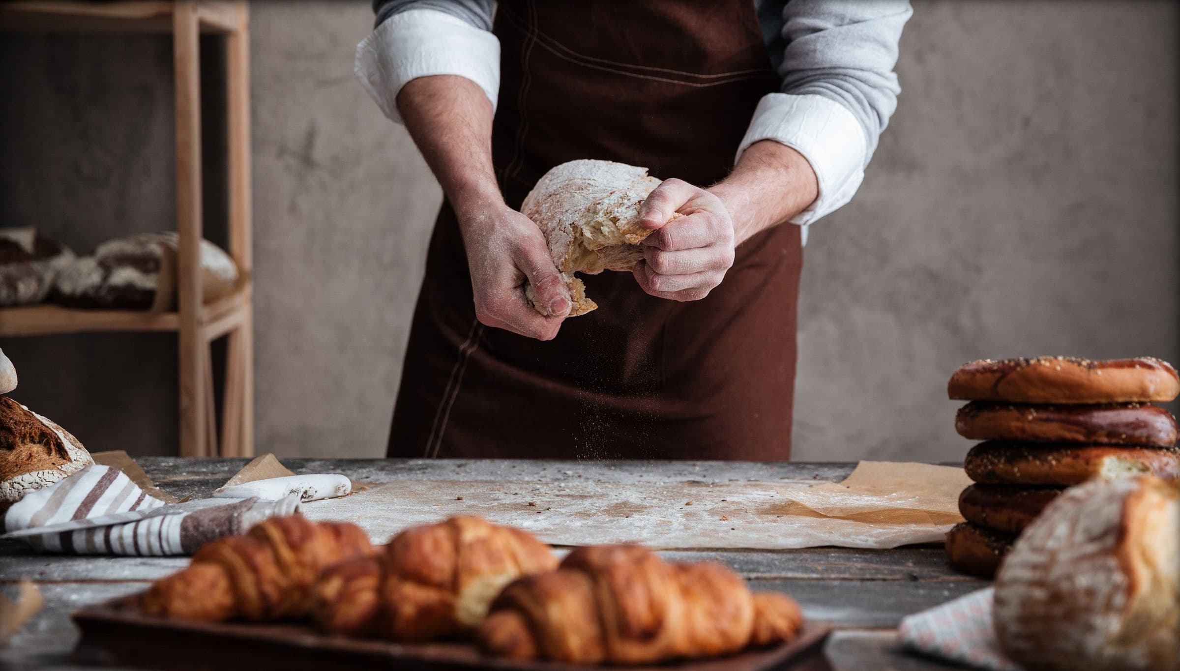 Baker in a brown apron tearing a floury loaf above a worktable lined with croissants and stacked bagels