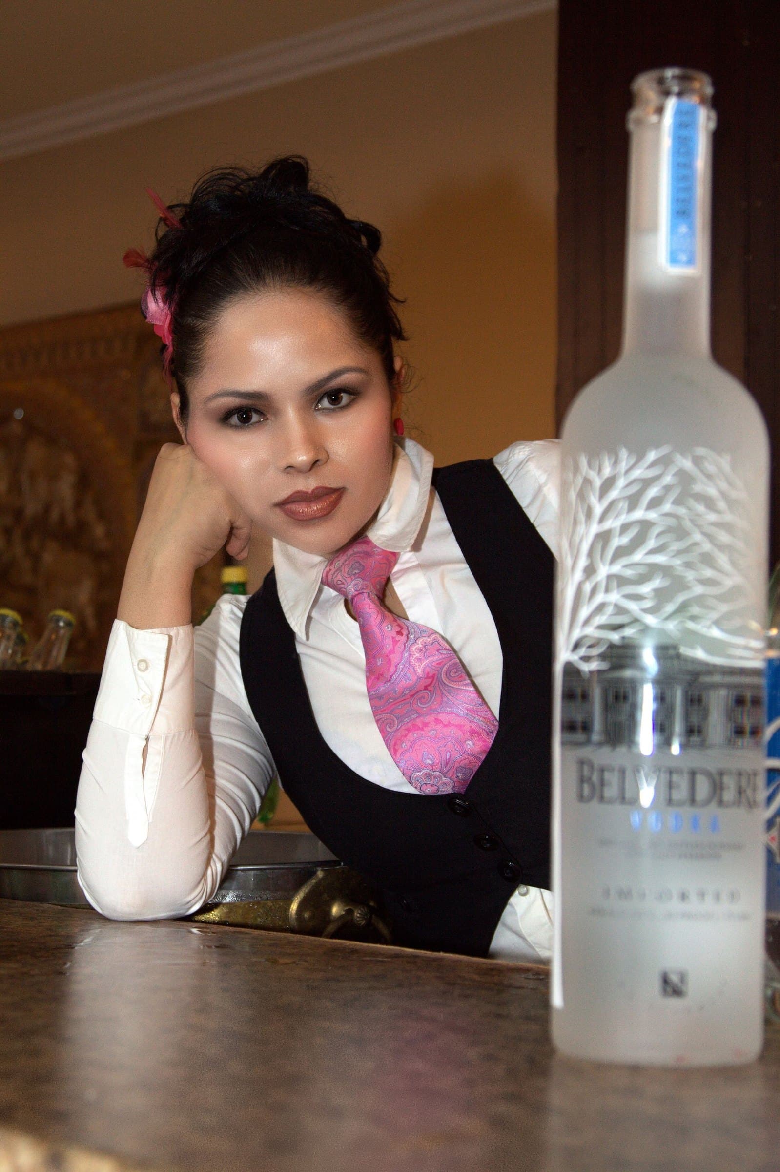 Bartender in a black waistcoat and pink tie leaning on the bar beside a frosted bottle of vodka