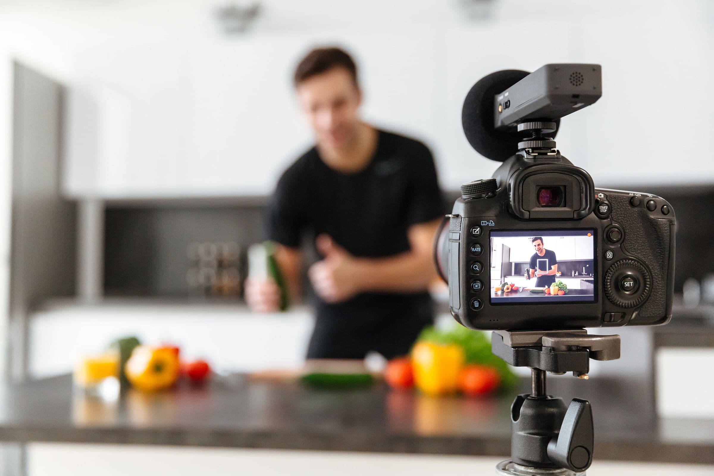 Camera on a tripod filming a smiling man preparing fresh vegetables in a bright kitchen, the shot visible on its screen