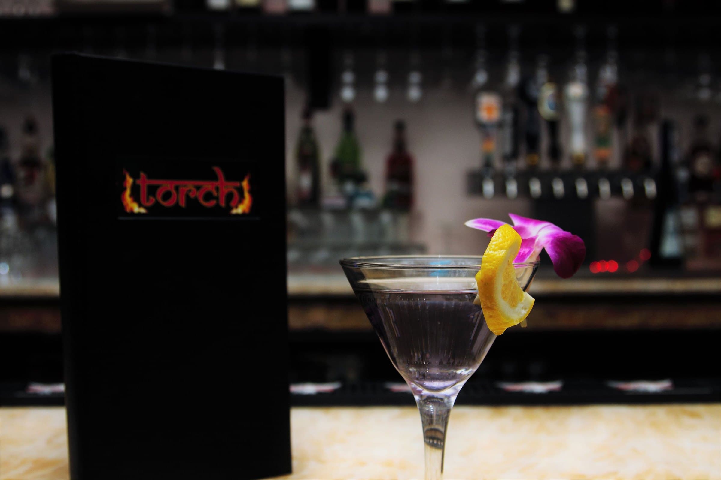 Martini garnished with a lemon twist and an orchid on a bar counter beside a black menu, shelves of bottles blurred behind