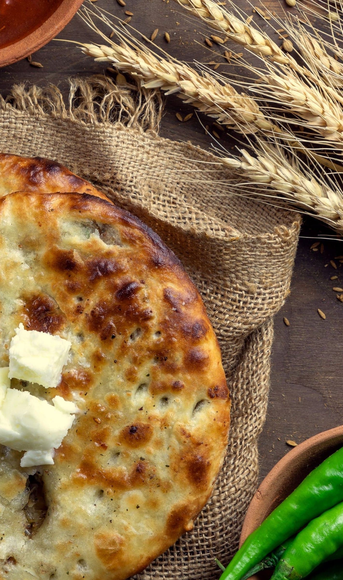 Charred naan topped with cubes of butter on a burlap cloth, with wheat stalks and green chillies around it