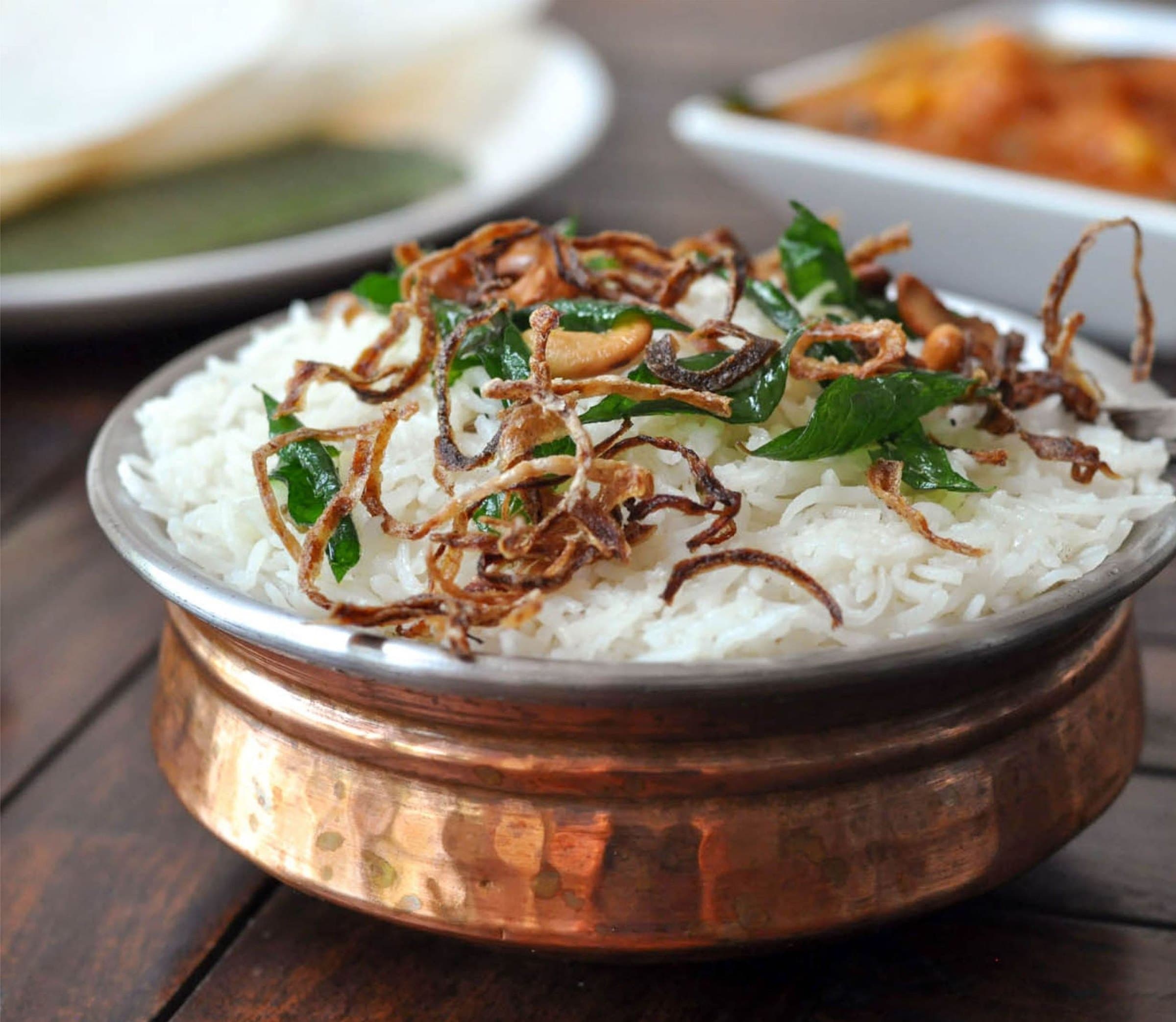 Steamed rice topped with crispy fried onions and curry leaves in a hammered copper bowl, curry and chutney dishes behind