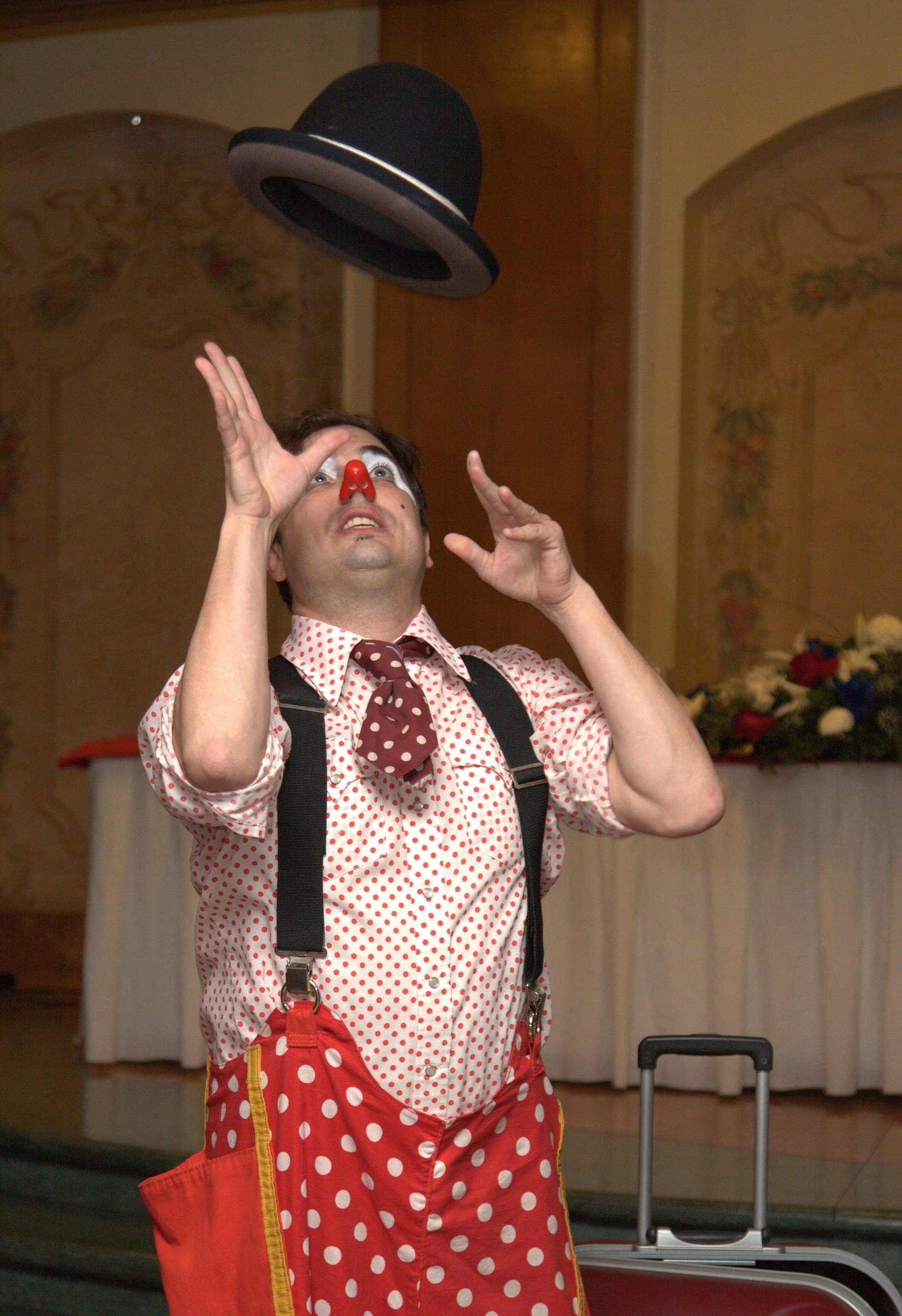 Entertainer in clown make-up, red nose and polka-dot shirt tossing a black bowler hat above his head in a banquet hall