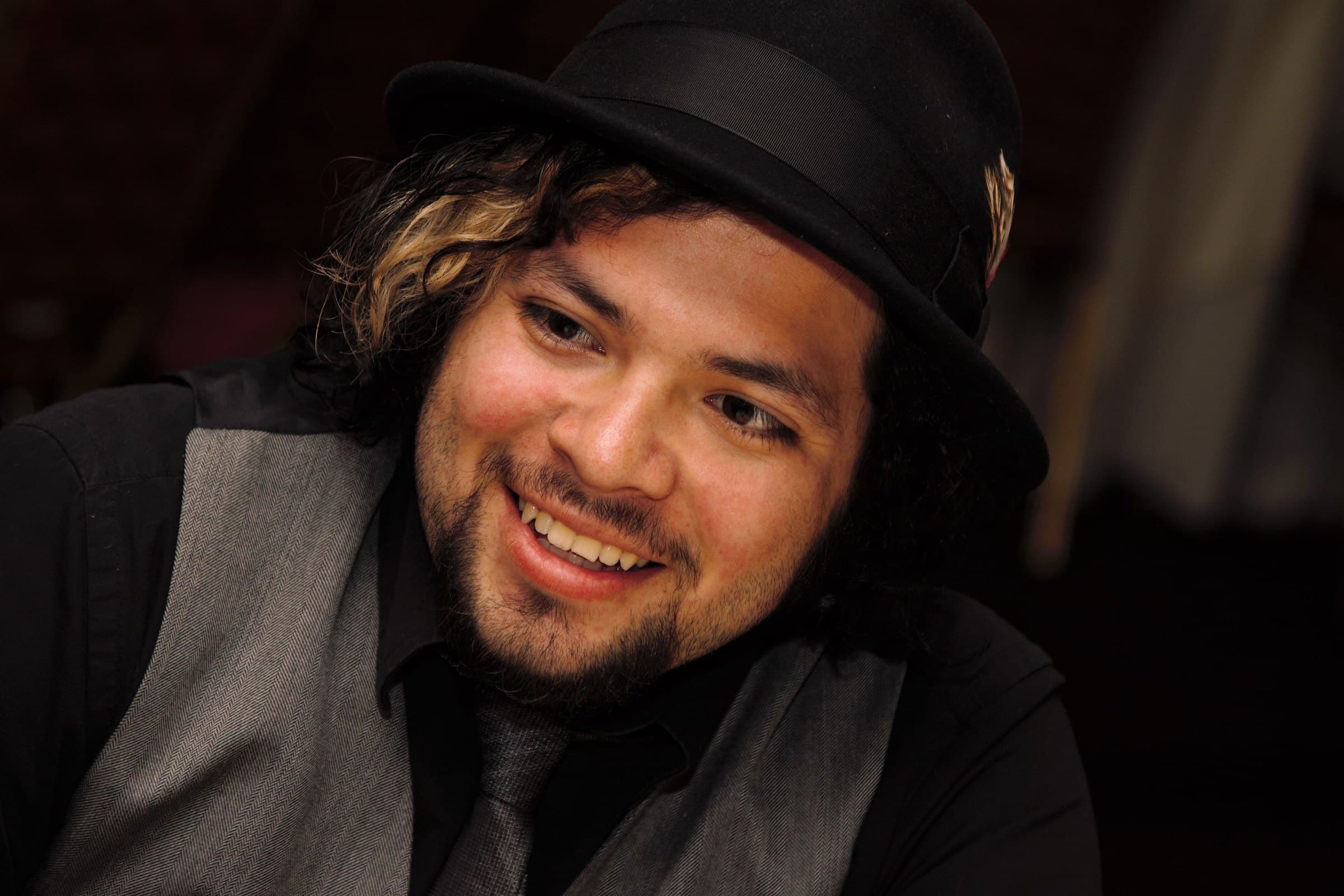 Man in a black bowler hat and grey waistcoat laughing as he glances to the side in a dimly lit venue