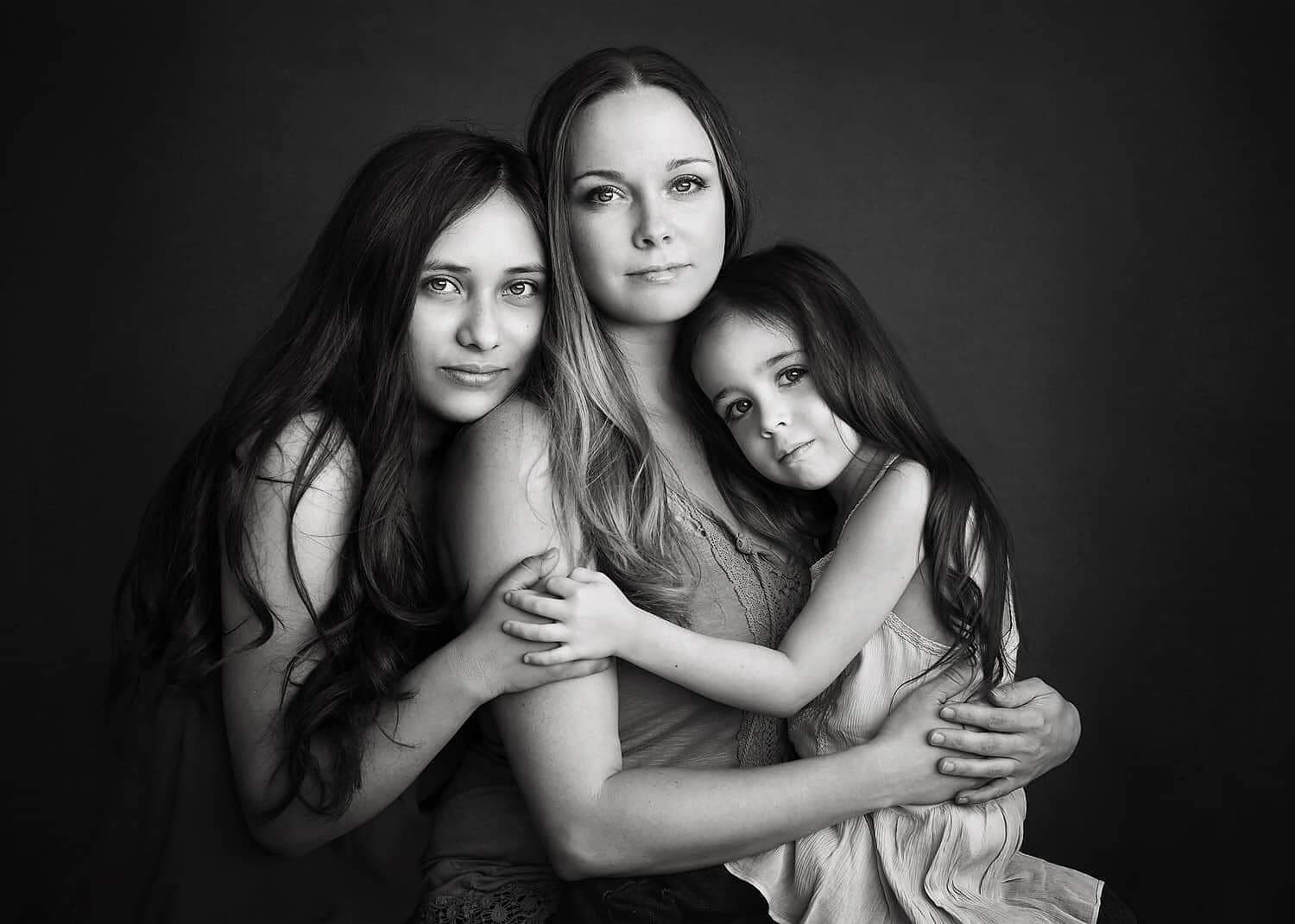 Black-and-white studio portrait of a mother embracing her two daughters, all three looking toward the camera