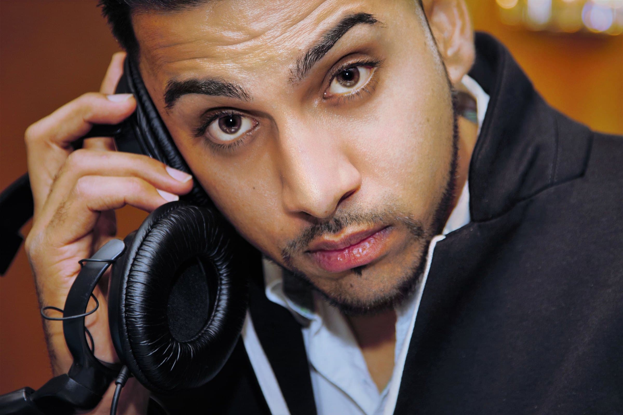 Young man in a dark jacket holding studio headphones to one ear, looking straight into the camera against a warm backdrop