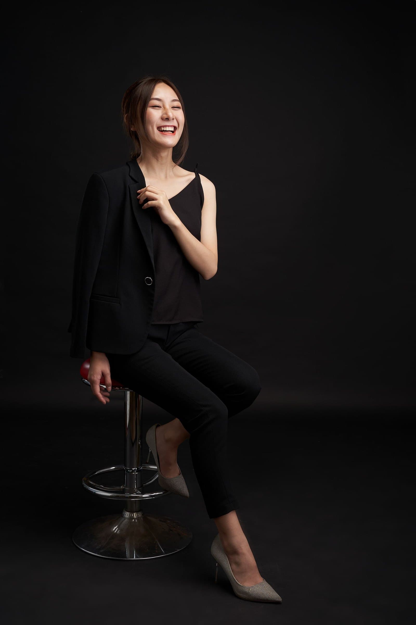 Woman in a black blazer laughing as she sits on a bar stool against a dark studio backdrop