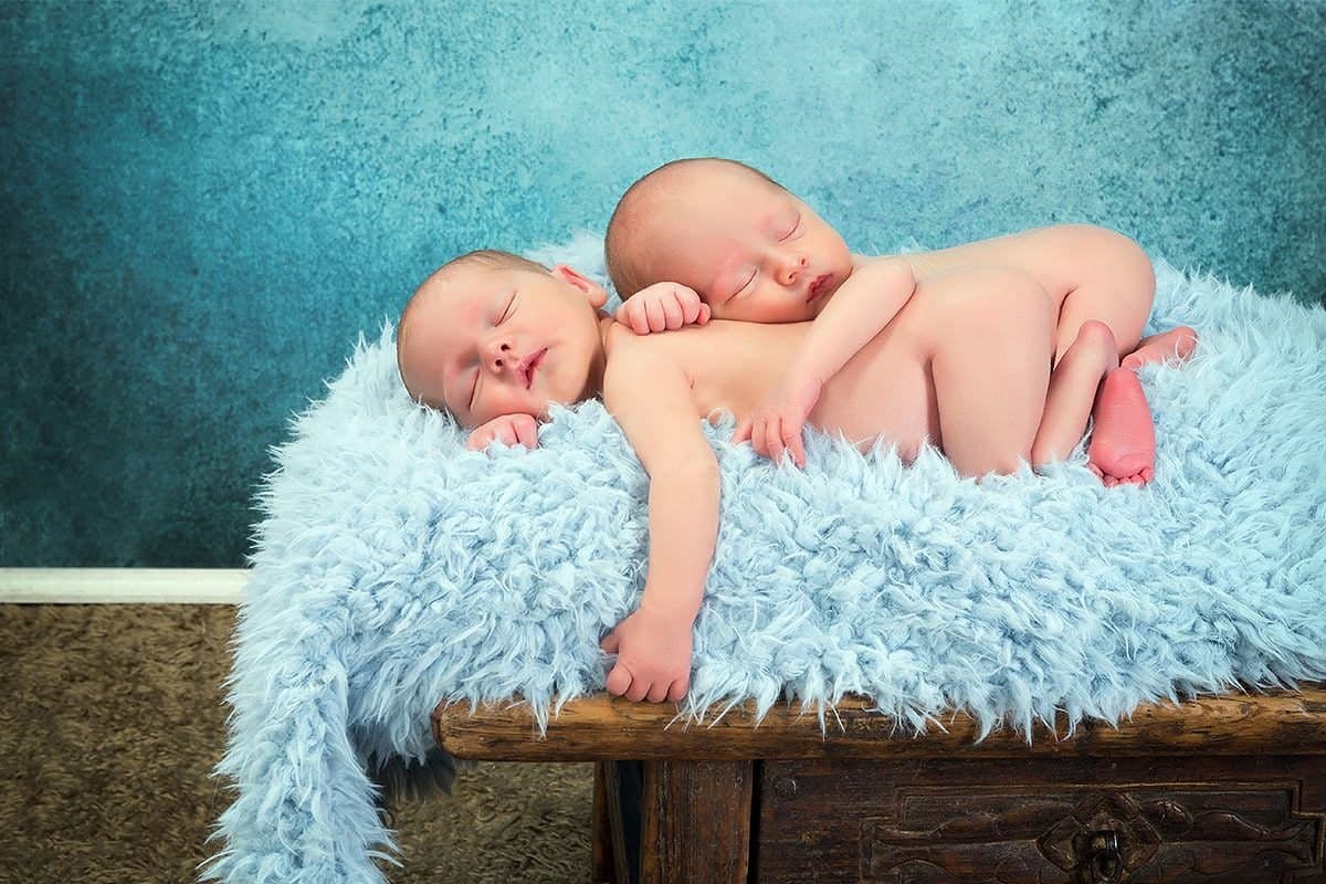 Newborn twins sleeping side by side on a fluffy blue blanket draped over a rustic wooden bench