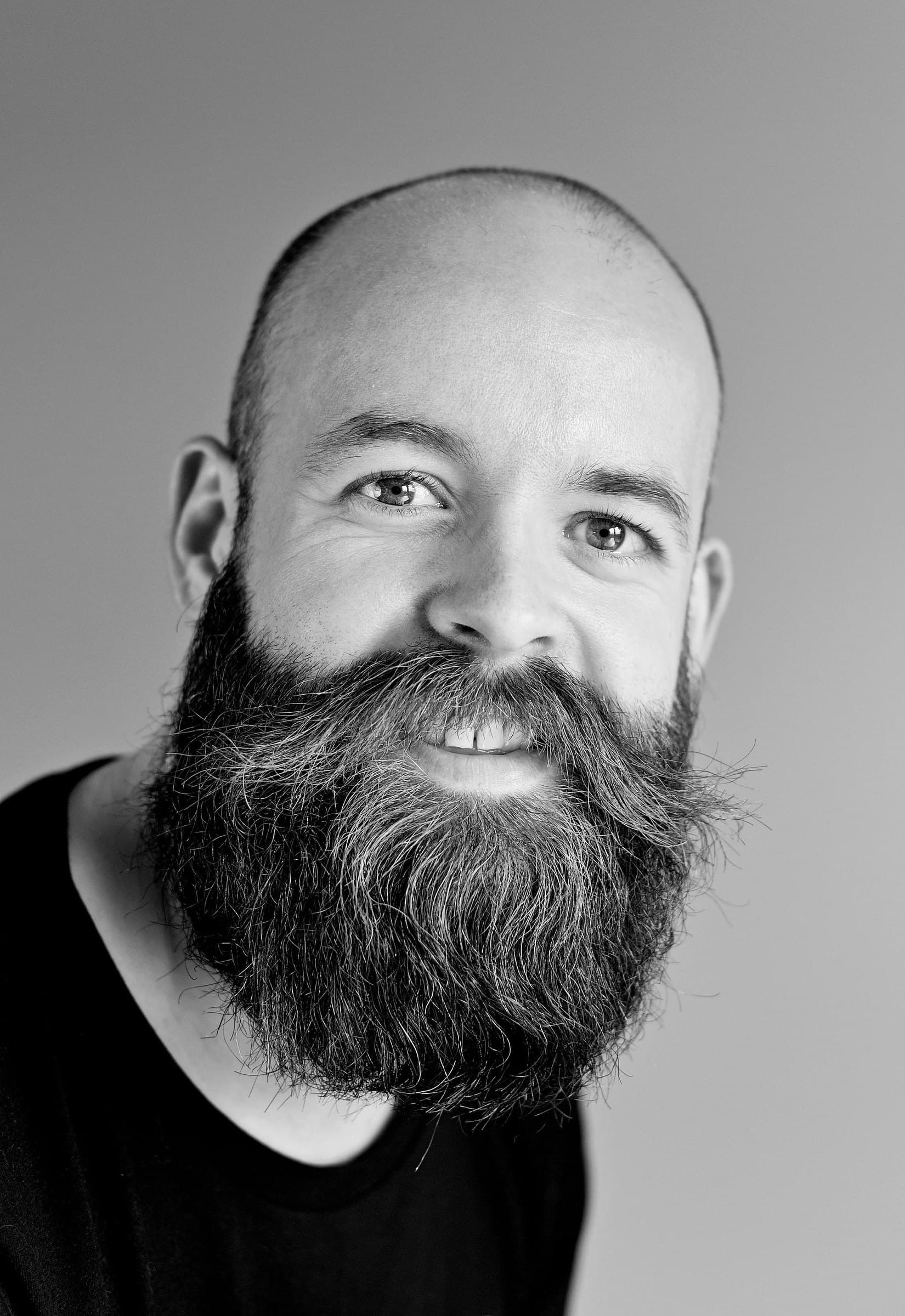 Black-and-white portrait of a smiling bald man with a full, long beard against a light grey backdrop
