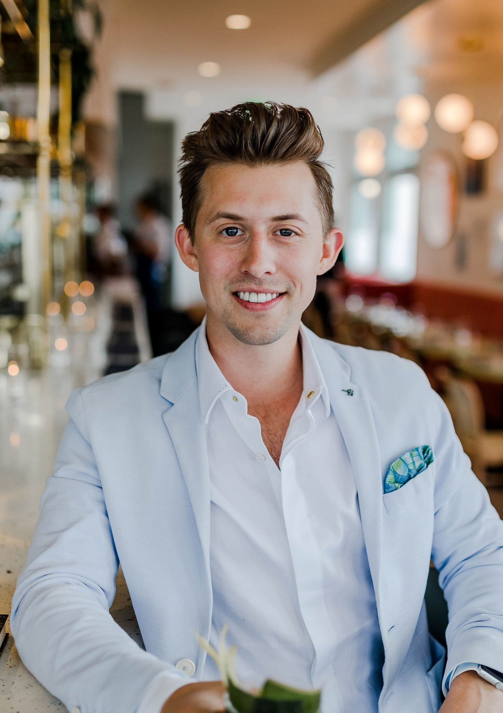 Smiling young man in a light blue blazer seated at a restaurant table, the dining room softly blurred behind him