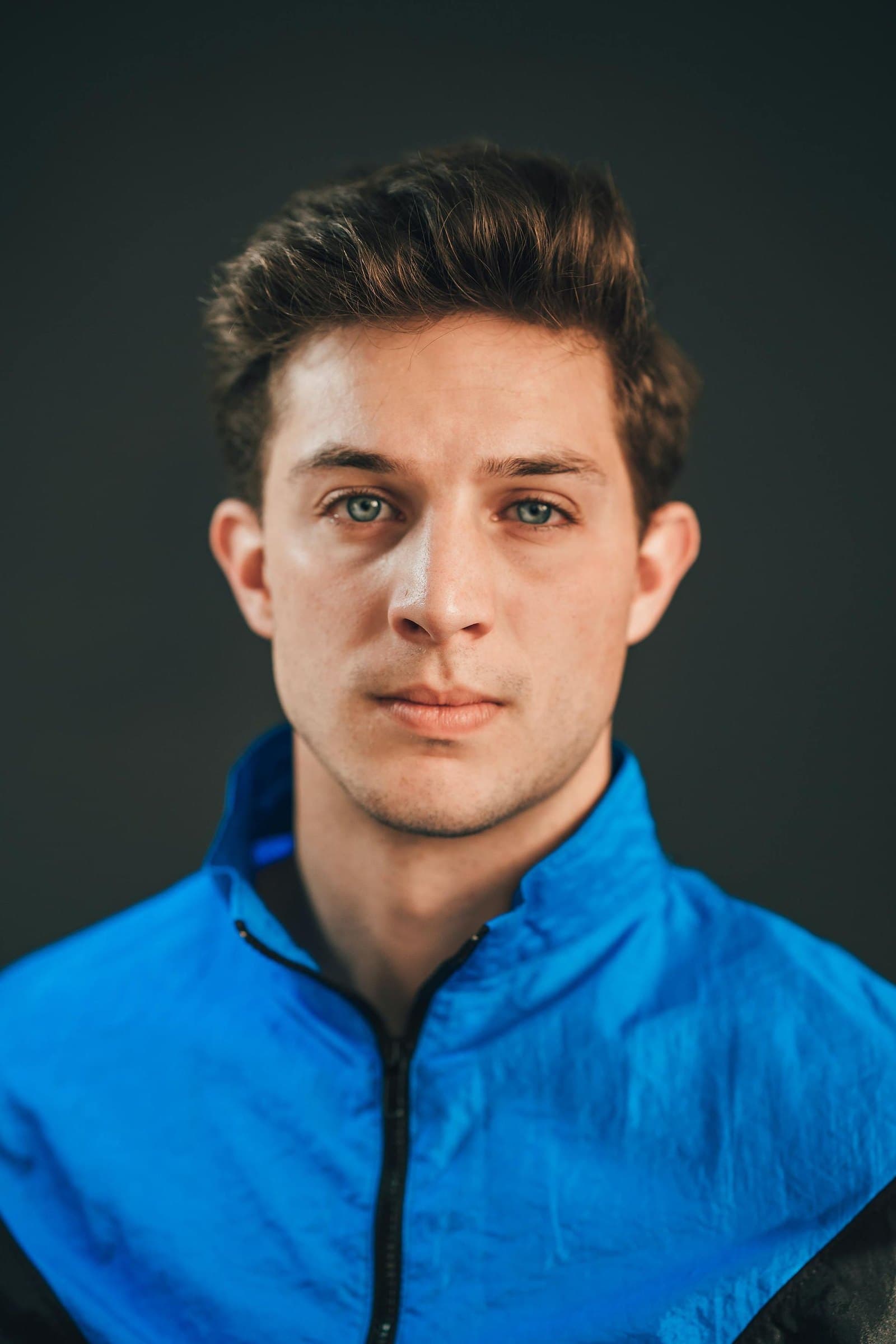 Young man with tousled brown hair in a blue zip-up jacket looking straight at the camera against a dark teal backdrop
