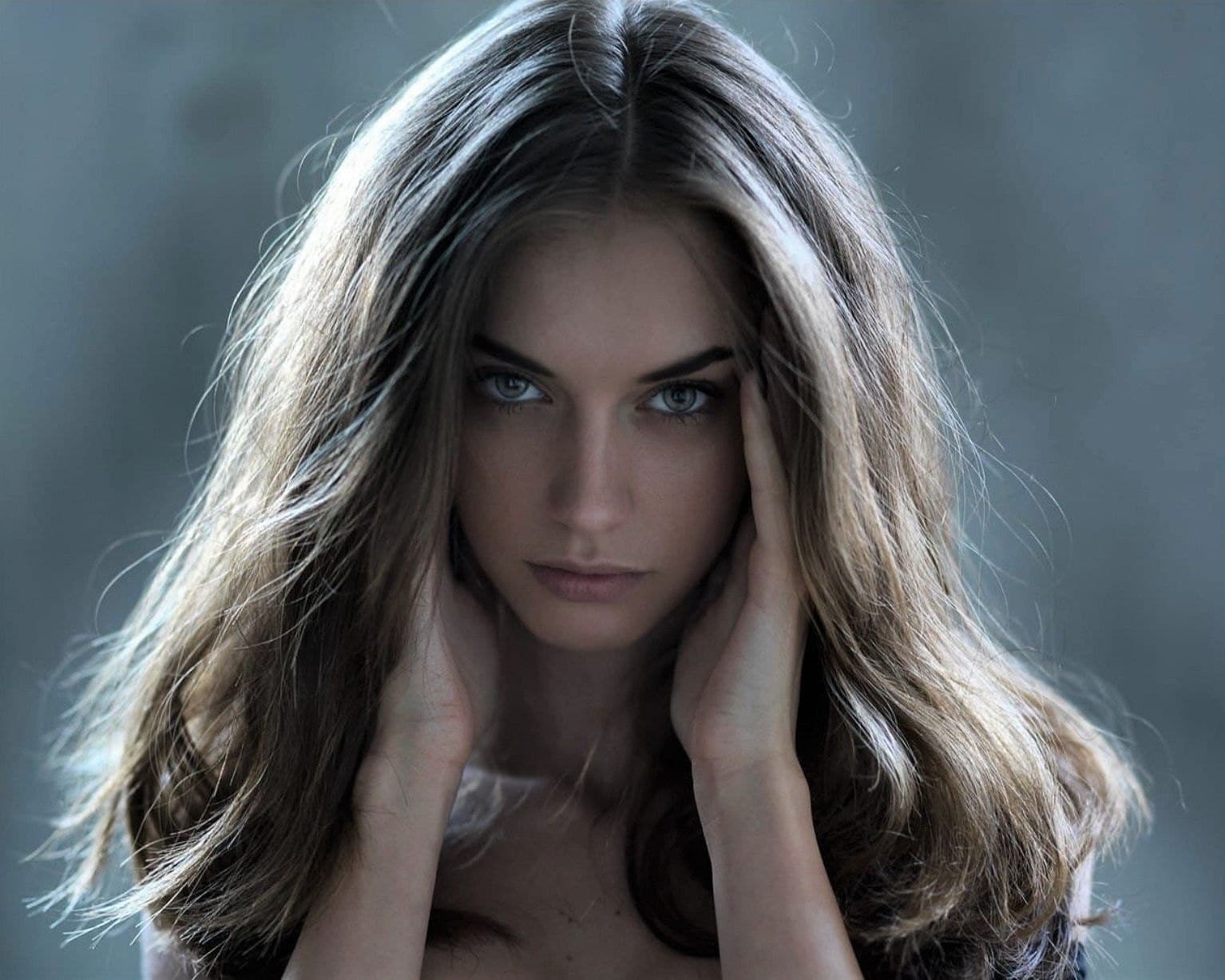 Woman with long tousled brown hair and pale eyes, hands raised to her face, against a soft blue-grey background