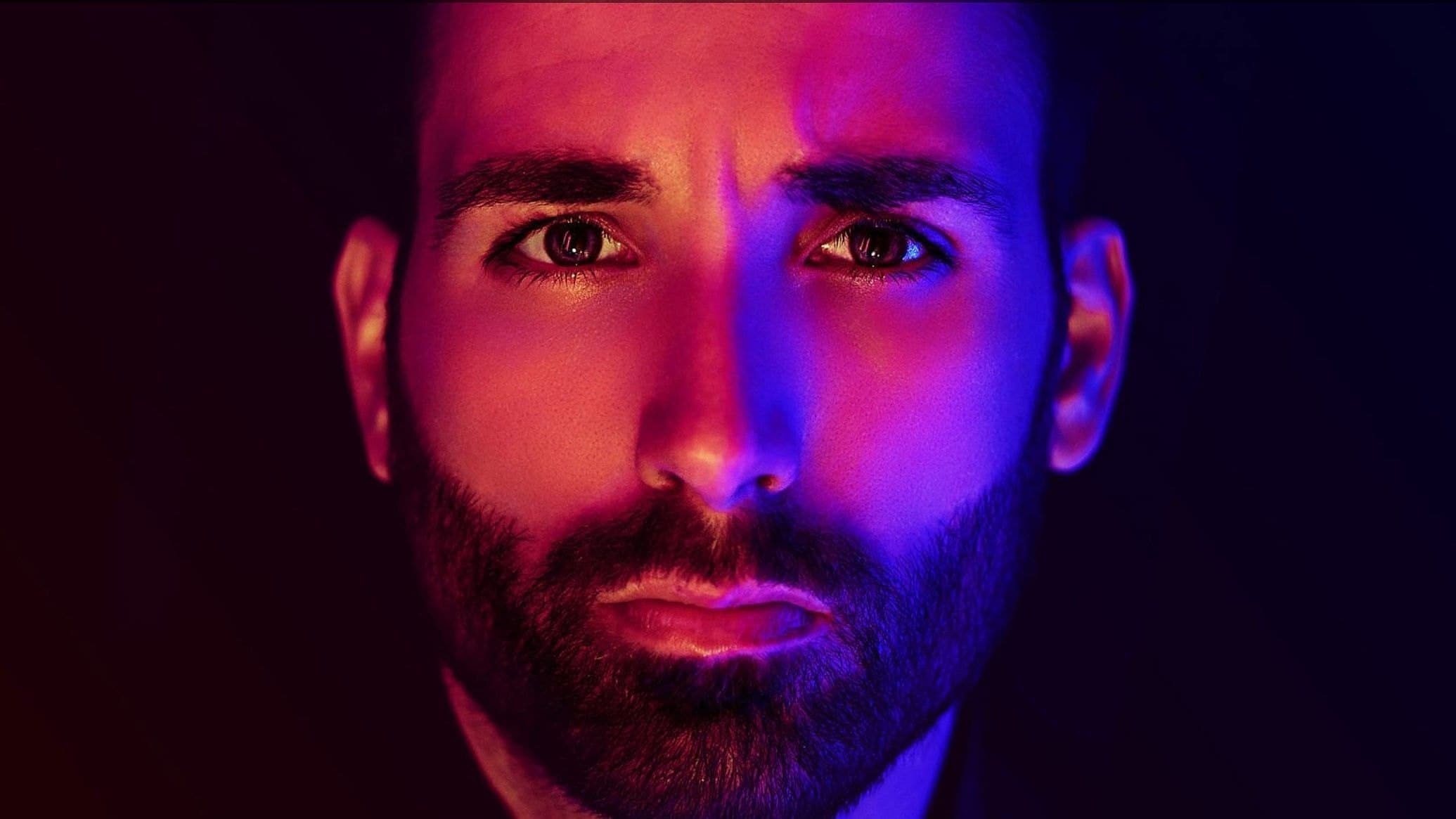 Bearded man with a steady expression lit in saturated magenta and blue light against a dark background