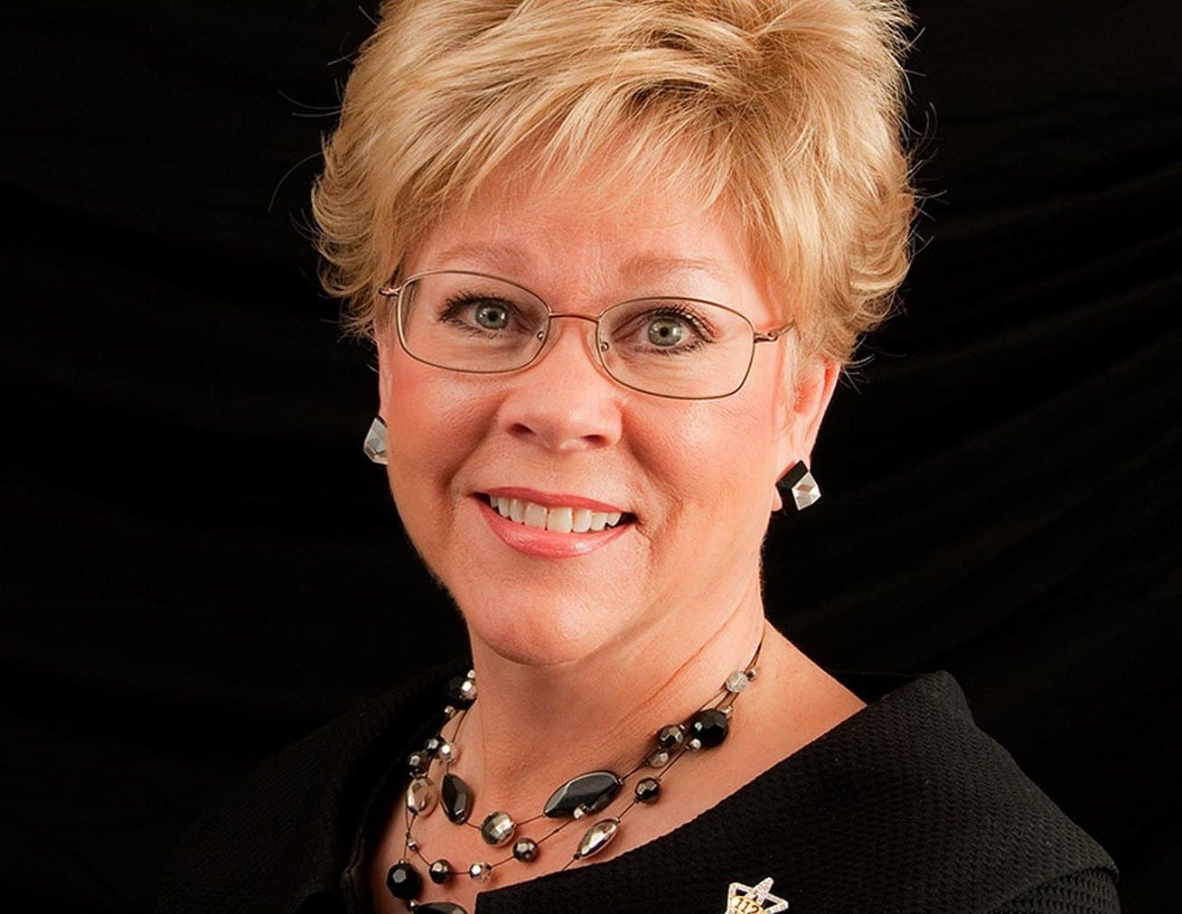Professional headshot of a smiling woman with short blonde hair, glasses and a beaded necklace against a black backdrop