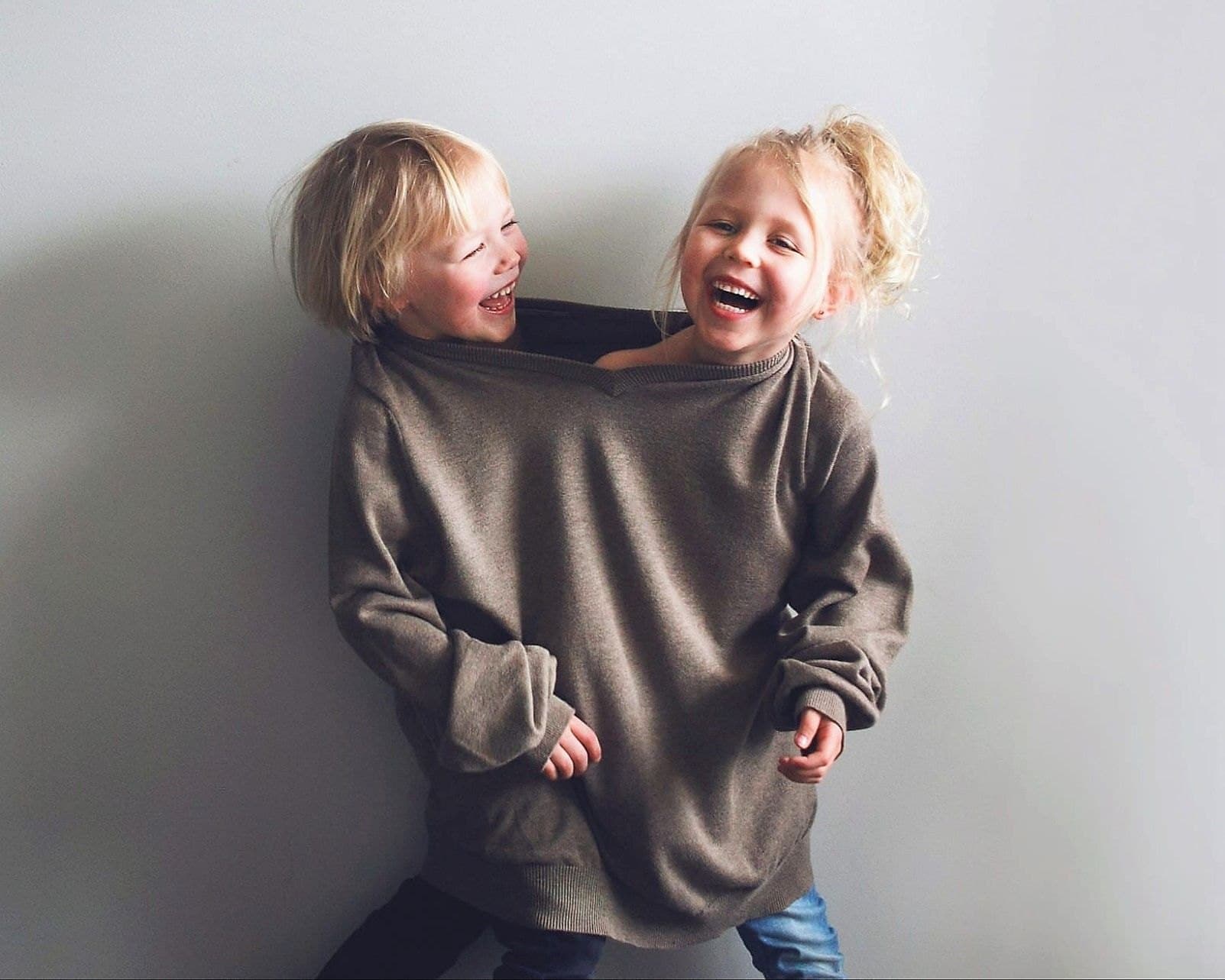 Two laughing blonde toddlers squeezed together inside one oversized grey sweater against a white wall