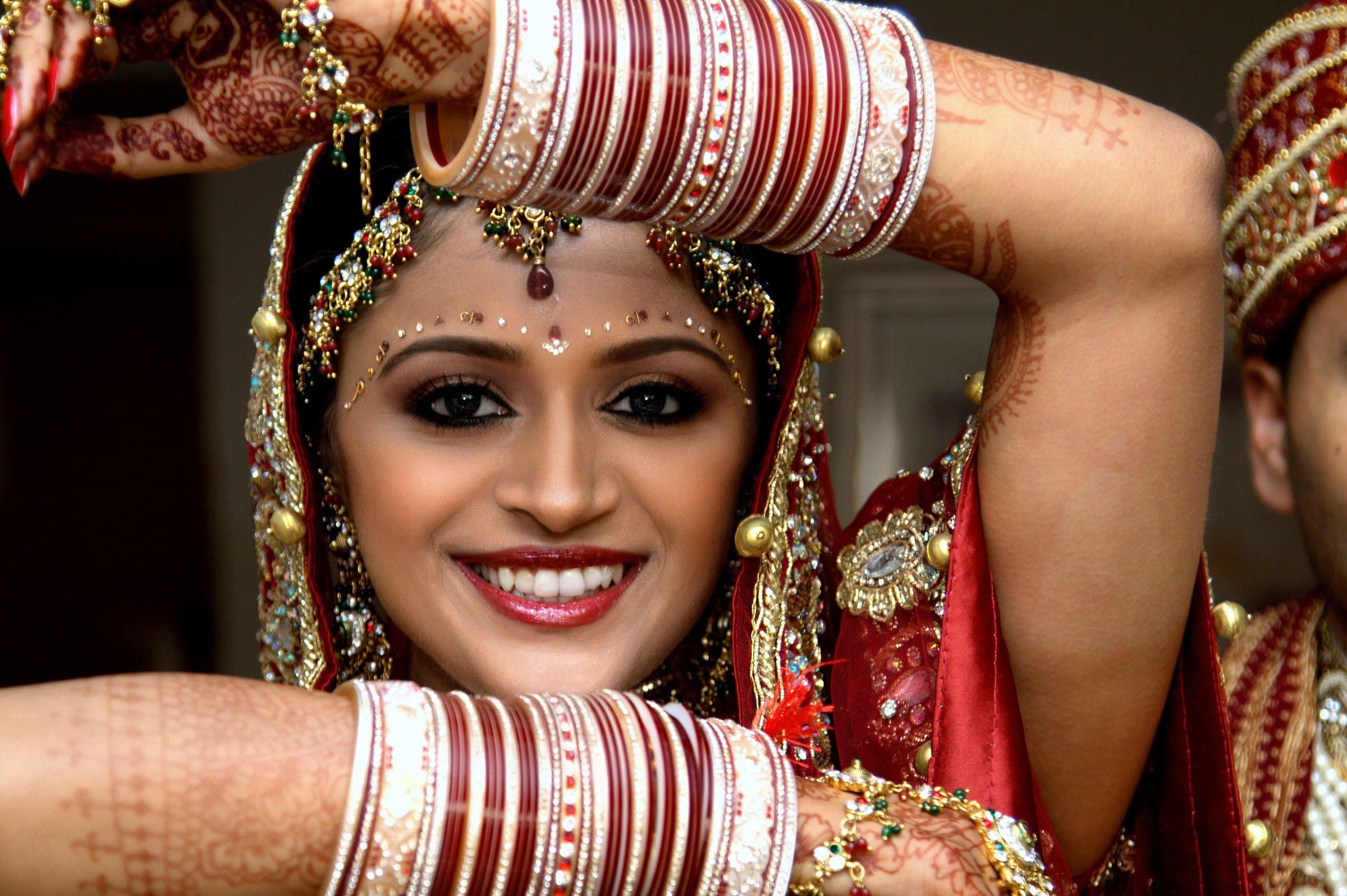 Smiling bride framing her face with her arms, stacked red and white wedding bangles and hennaed hands above her jewelled forehead