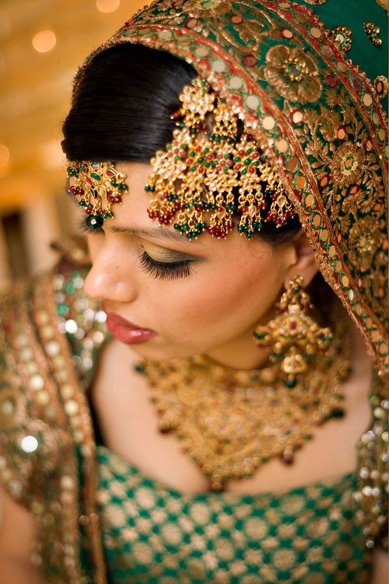 Profile of a bride looking down, an ornate gold and emerald headpiece and heavy earrings over a green lehenga