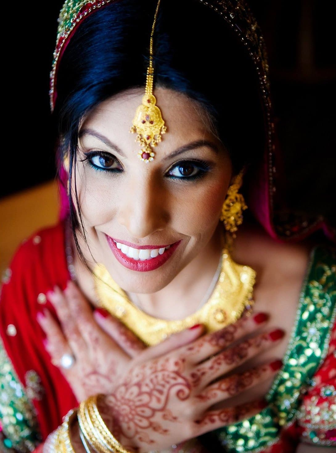 Bride with a gold maang tikka and necklace smiling up at the camera, her hennaed hand resting on her chest