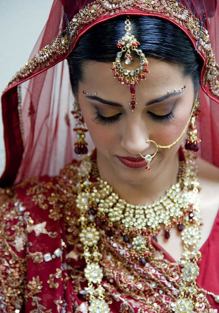 Bride in a red veil with a nose ring and layered kundan necklaces, eyes lowered in a quiet moment