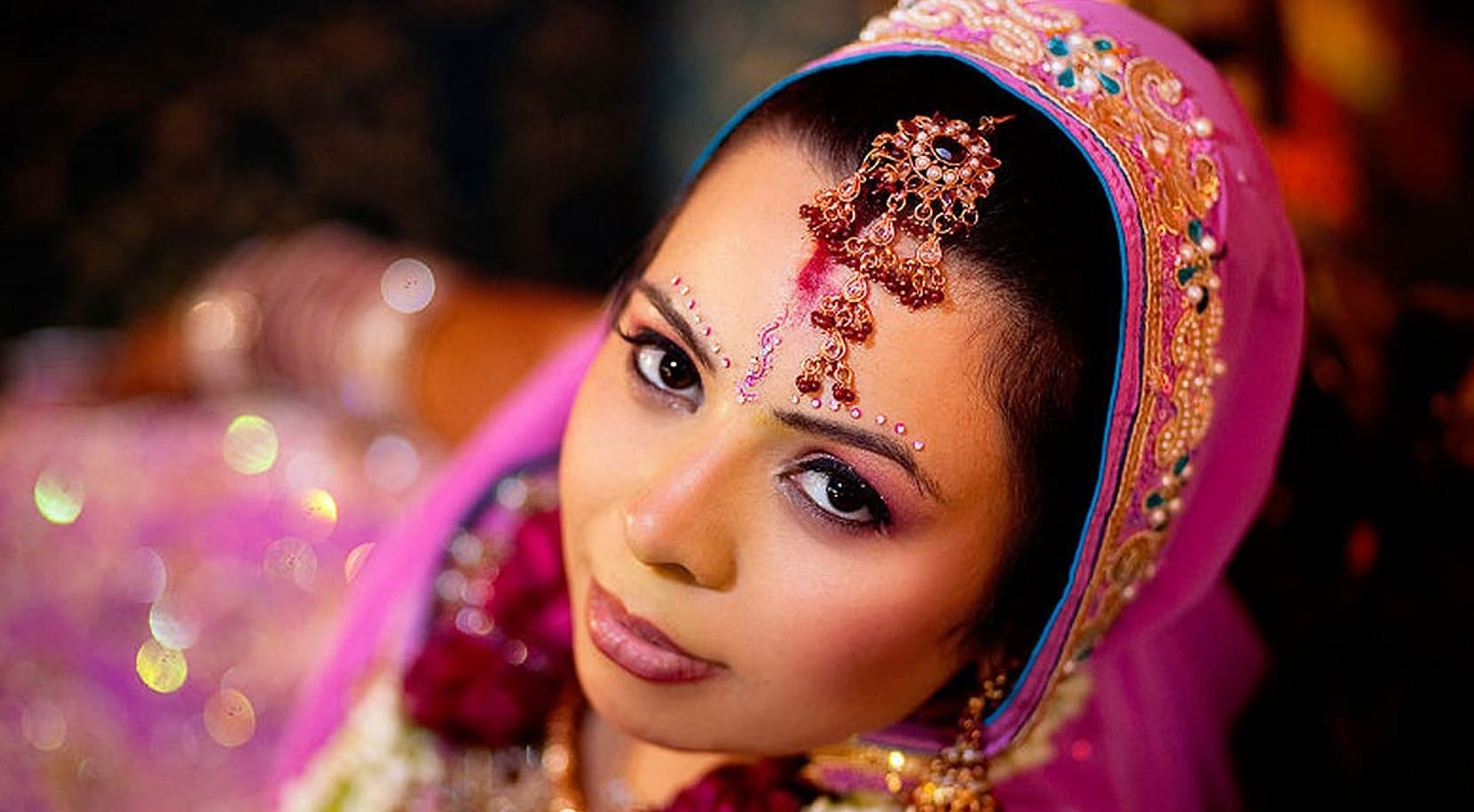 Bride in a pink embroidered dupatta tilting her head toward the camera, golden lights sparkling in the background