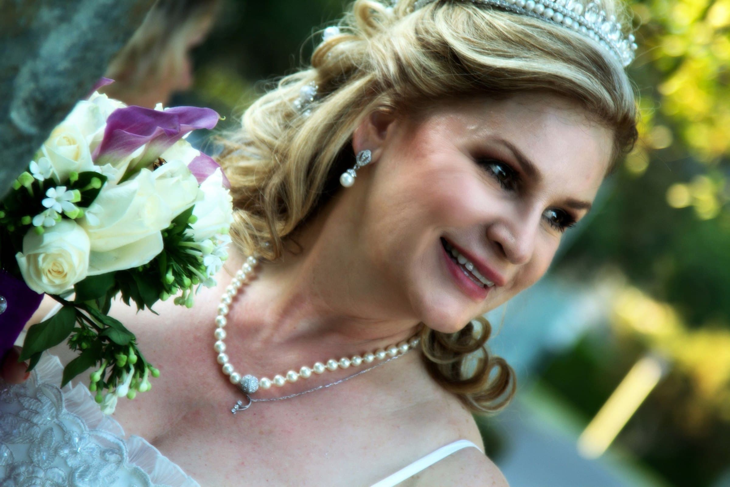 Bride in a tiara and pearl necklace smiling over her shoulder beside a bouquet of white roses and purple calla lilies