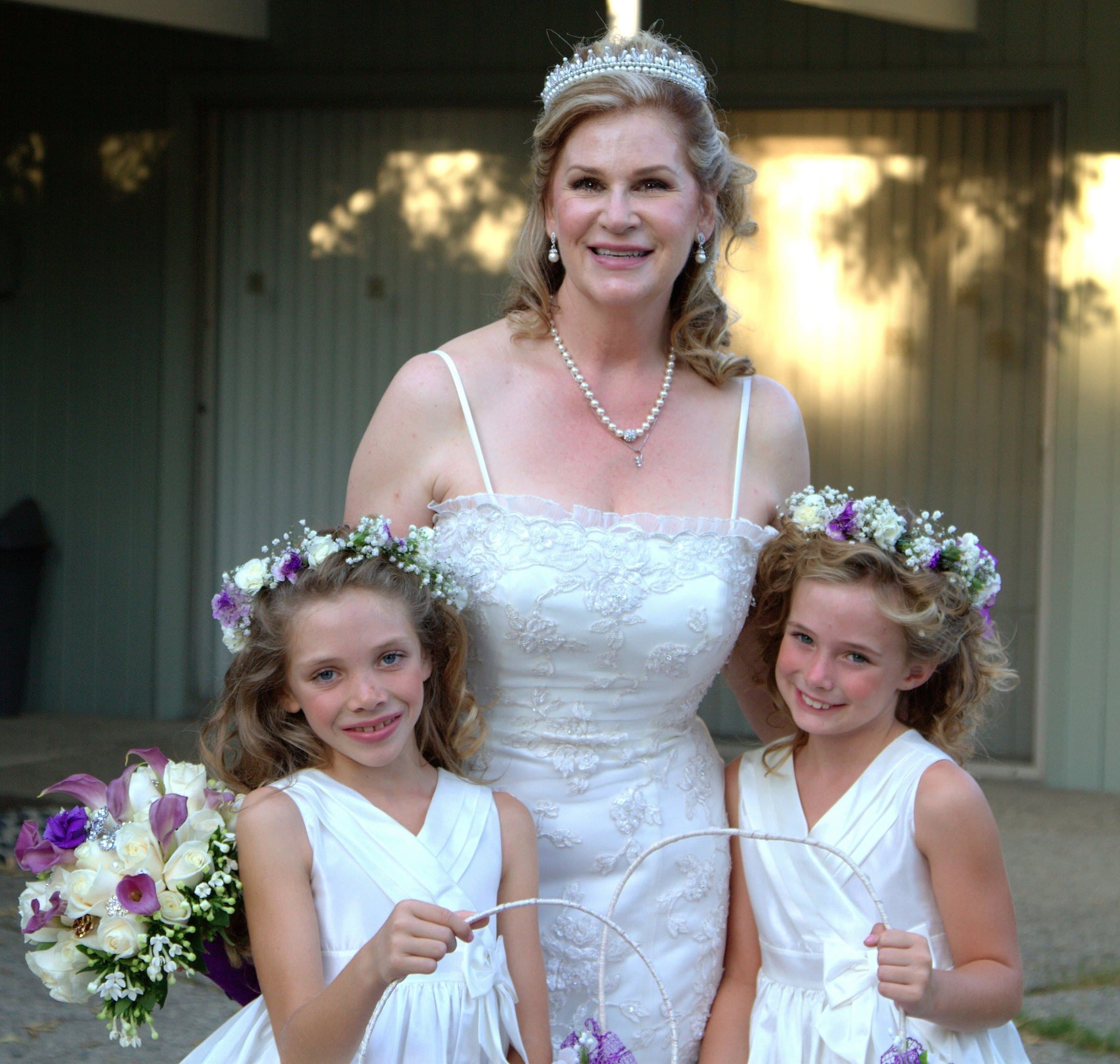 Bride in a white gown and tiara standing between two smiling flower girls wearing floral crowns and holding baskets