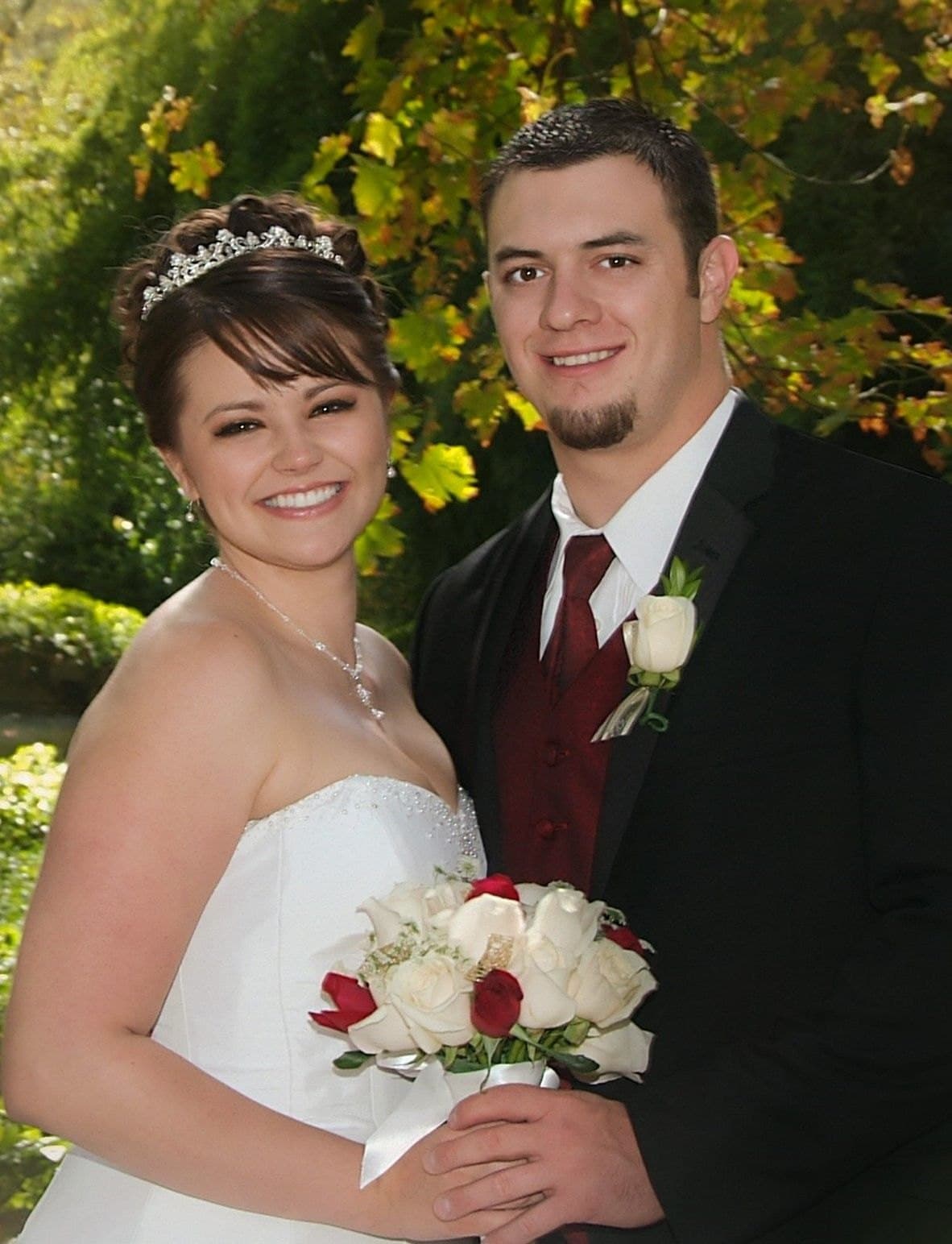Smiling bride and groom posed close together in front of autumn leaves, the bride holding white and red roses